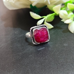 Puede incluir: Un anillo de plata con una piedra preciosa de color rojo intenso, tallada en forma cuadrada y facetada, engastada en un marco de metal texturizado. El aro es liso y pulido, contrastando con los detalles oscuros y complejos alrededor del engaste.