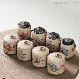 May include: A selection of ceramic jars with lids and floral and botanical designs. The jars are beige with gold ring handles. Some jars have fabric tops with floral patterns. The text "tsukiceramics" is in the upper right corner.