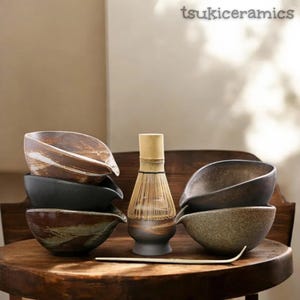 May include: A selection of ceramic matcha bowls in earthy tones, arranged on a wooden table. A bamboo whisk and a scoop are also included. The bowls feature a distinctive spout design. The text "tsukiceramics" is visible in the top right corner.