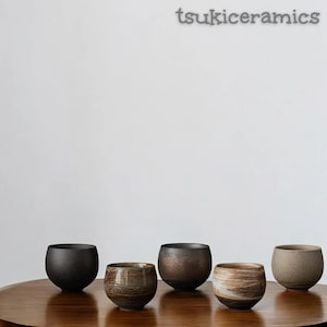 May include: Five ceramic bowls in shades of black, brown, and beige are displayed on a wooden table. The bowls have a rounded shape and textured surfaces, showcasing the unique characteristics of the pottery. The brand name "tsukiceramics" is visible.