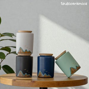 May include: Four ceramic jars with wooden lids, each decorated with a mountain and bird motif. The jars are in white, black, blue, and mint green. The brand name "tsukiceramics" is visible in the top right corner.