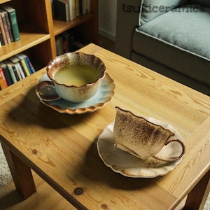 May include: Two ceramic teacups and saucers on a wooden table. The cups have a unique, textured design with brown and cream tones, and the saucers are light blue with a scalloped edge. One cup is filled with a light green liquid.