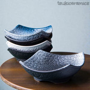 May include: A stack of square ceramic bowls in various colors: blue, black, white, and gray. The bowls have a speckled texture and a unique, wavy rim design. The bowls are on a wooden surface.