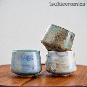 May include: Three handmade ceramic cups with a unique, textured glaze. The cups are in shades of blue, green, and brown, stacked on a wooden surface. The text "tsukiceramics" is in the upper right corner.