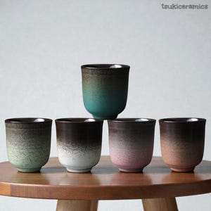 Puede incluir: Cinco tazas de té de cerámica con un diseño degradado moteado. Las tazas son en tonos de verde, marrón, rosa y beige, con un borde marrón oscuro. Las tazas están dispuestas sobre una mesa de madera. El texto "tsukiceramics" está en la esquina superior derecha.
