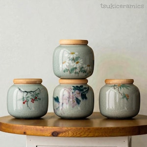 Può includere: Quattro barattoli in ceramica con coperchi in legno. I barattoli sono di colore verde chiaro e presentano disegni floreali dipinti a mano in bianco, rosso e viola. I barattoli sono impilati su una superficie di legno. Il testo "tsukiceramics" è in alto a destra.