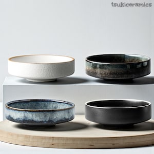 Puede incluir: Cuatro cuencos de cerámica en varios colores: blanco con borde marrón, verde oscuro y marrón, azul con detalles blancos y negro. Los cuencos son bajos y anchos, adecuados para servir. La marca "tsukiceramics" está en la esquina superior derecha.