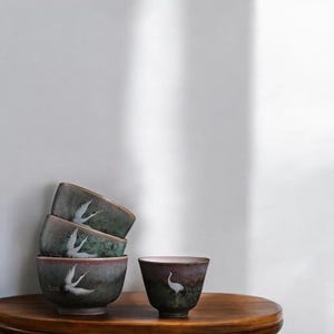 May include: Four ceramic tea bowls with a crane design. The bowls are stacked on a wooden table. The bowls have a gradient colour scheme, transitioning from a dark maroon to a teal green. One bowl is separate, showing the crane design.