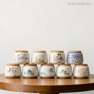 Puede incluir: Una colección de tarros de cerámica con tapas de madera. Cada tarro presenta un diseño único pintado a mano, incluyendo paisajes, patrones florales y un gato. Los tarros están dispuestos sobre una superficie de madera, mostrando su atractivo decorativo.