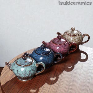 Può includere: Quattro teiere in ceramica in fila, ognuna con un colore e un motivo unici. Le teiere sono in tonalità di verde acqua, blu, rosa e beige, con accenti marroni. Le teiere sono su una superficie di legno. Il testo "tsukiceramics" è in alto a destra.