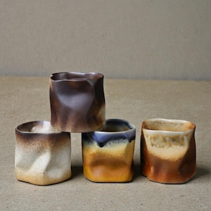 May include: Four handcrafted ceramic cups with a unique, textured design. The cups feature a square shape with folded sides and a variety of earthy tones, including brown, beige, and yellow. The cups are stacked, showcasing their individual colors and textures.