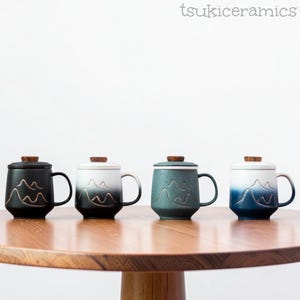 May include: Four ceramic mugs with lids, each featuring a mountain range design. The mugs are black, white to black gradient, teal, and blue to white gradient. Each mug has a wooden lid and a black handle. The mugs are displayed on a wooden table.