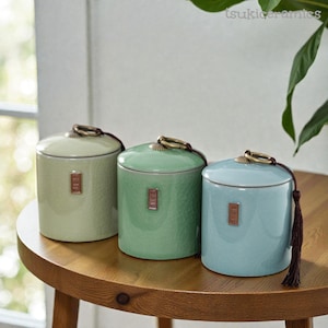 Op de afbeelding: Drie keramische potten in pastelgroen, groen en blauw, elk met een deksel en een decoratieve metalen ringhandgreep. De potten hebben een gestructureerd oppervlak en een klein rechthoekig embleem aan de voorkant. Een bruine kwast hangt aan de blauwe pot.