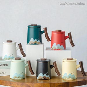 May include: A selection of ceramic mugs with lids and wooden handles. Each mug features a mountain landscape design in various colours, including white, teal, red, black, and yellow. The mugs are displayed on a wooden surface and white pedestals.