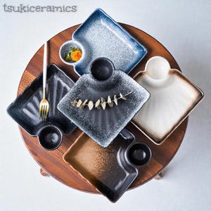 May include: A selection of square ceramic plates and small bowls in shades of black, blue, and brown, displayed on a wooden surface. Some plates feature an attached bowl. A gold fork is also visible. The plates are arranged in a circular pattern.