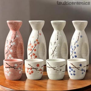 May include: A ceramic sake set featuring four bottles and three cups. The bottles are white and pink, decorated with floral branch designs. The cups have matching designs. The set is displayed on a wooden surface.