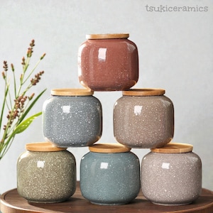 May include: A stack of six ceramic jars in various colors, including gray, brown, and blue, each with a speckled finish and a natural wood lid. The jars are arranged in a pyramid shape. The brand name "tsukiceramics" is in the top right corner.