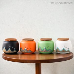 May include: Four ceramic jars with wooden lids, each painted a different color: black, orange, green, and white. Each jar features a mountain and bird design in shades of blue, brown, and white. The jars are displayed on a round wooden table.