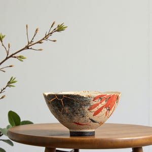 May include: A ceramic bowl with a distinctive design. The bowl has a black and blue section with gold detailing, and a section with red leaf patterns. The bowl is on a wooden table, with a branch in the background.