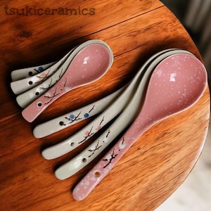 Könnte beinhalten: Eine Sammlung von Keramik-Löffeln mit rosa gesprenkelten Schalen und weißen Griffen. Die Griffe sind mit einem floralen Design in Schwarz, Blau und Rosa verziert. Die Löffel sind auf einer Holzoberfläche angeordnet. Der Text "tsukiceramics" ist sichtbar.
