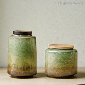 May include: Two ceramic jars with wooden lids. The jars have a gradient glaze, transitioning from green to brown. The larger jar is taller than the smaller one. The jars are cylindrical and sit on a light-coloured surface. The text "tsukiceramics" is in the top right corner.