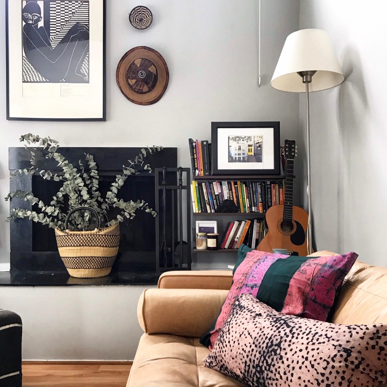 May include: A living room interior with a tan couch, a bookshelf with books and a guitar, a black fireplace, and a potted plant. The couch has two pillows, one with a pink and green pattern and the other with a black and white spotted pattern.