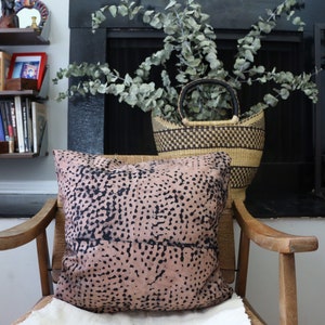 May include: A wooden armchair with a pink and black patterned pillow. The chair is in front of a fireplace with a black basket filled with eucalyptus branches.