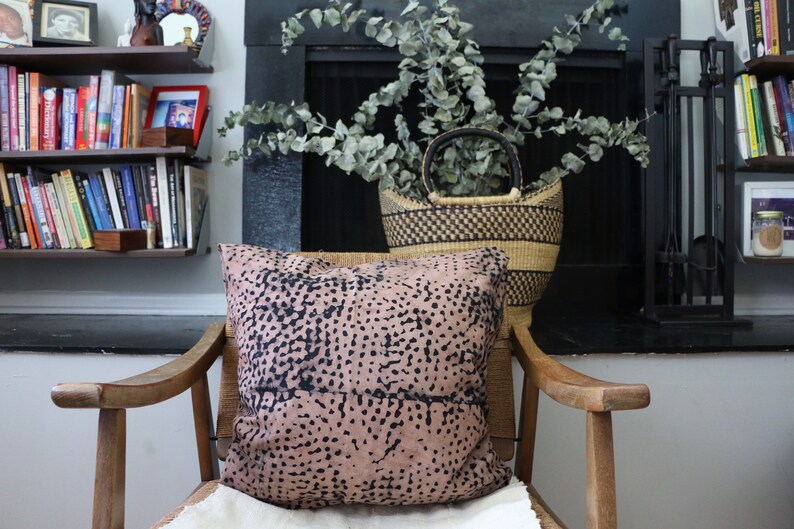 May include: A wooden armchair with a pink and black patterned pillow. The chair is in front of a fireplace with a black basket filled with eucalyptus branches.