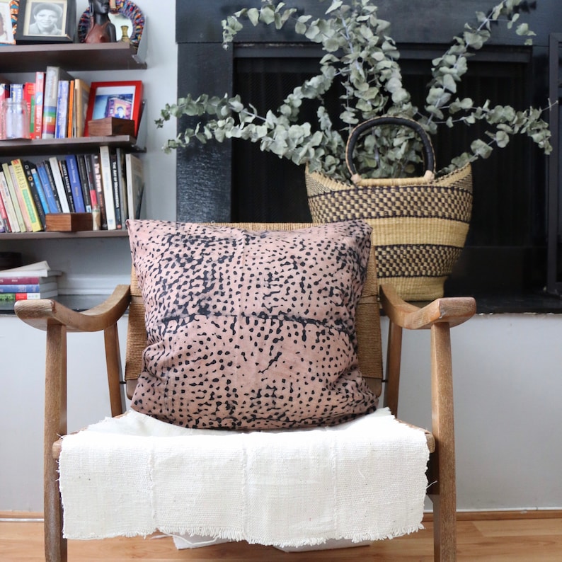 May include: A wooden chair with a white throw blanket and a pink and black patterned pillow. The chair is in front of a fireplace with a basket of greenery.