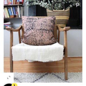 May include: A wooden chair with a white throw blanket and a pink and black patterned pillow. The chair is in front of a bookshelf and a basket.
