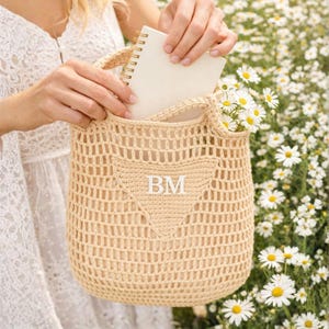 Puede incluir: Un bolso tote de crochet beige con un diseño triangular y las iniciales "BM" en blanco. El bolso contiene un cuaderno con espiral y un ramo de margaritas blancas con centros amarillos. El bolso es sostenido por una persona.