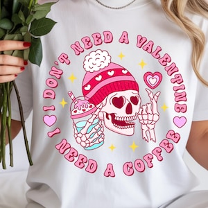 May include: White t-shirt with a graphic design featuring a skull wearing a pink beanie and holding a coffee drink. The text reads "I don't need a Valentine, I need a coffee."