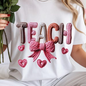 May include: White t-shirt featuring the word "TEACH" in a textured, 3D design. The letters are in shades of brown, pink, and orange, with a pink bow and heart accents. The shirt is part of a Valentine's Day theme.