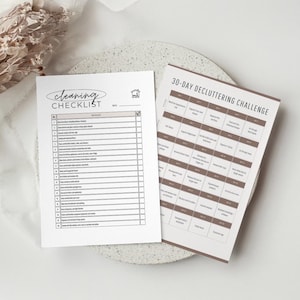 Op de afbeelding: Twee gedrukte checklists op een wit oppervlak. Eén is een "Cleaning Checklist" met schoonmaaktaken. De andere is een "30-Day Decluttering Challenge" met dagelijkse opruimopdrachten. Beide zijn in minimalistische stijl.
