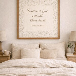 Puede incluir: Un cuadro enmarcado con el texto "Trust in the Lord with all thine heart" cuelga sobre una cama con ropa de cama beige. El marco es marr&oacute;n y el texto est&aacute; en una fuente de escritura. La cama tiene varias almohadas y dos l&aacute;mparas de noche.