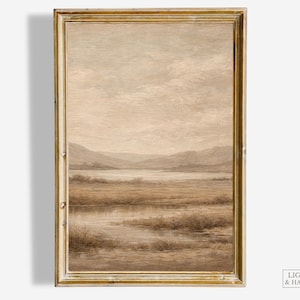 May include: A framed landscape art print featuring a serene marsh scene in muted tones of beige, brown, and gray. The artwork depicts a body of water, grassy areas, and distant hills under a cloudy sky. The frame is gold.