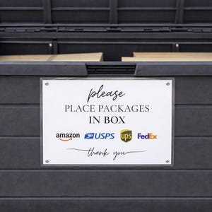 May include: A dark gray package drop box with an open lid. A white sign reads "Please Place Packages In Box" with logos for Amazon, USPS, UPS, and FedEx. The box is designed for secure package delivery.