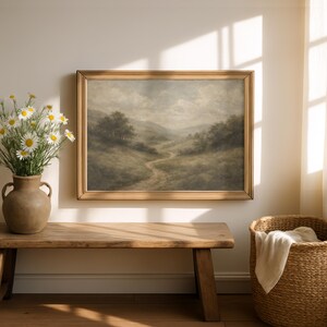Pode incluir: Uma pintura de paisagem emoldurada retrata um caminho sinuoso atrav&eacute;s de colinas sob um c&eacute;u nublado. A obra de arte est&aacute; em uma moldura dourada, pendurada acima de um banco de madeira. Um vaso de margaridas e uma cesta de vime completam a cena.