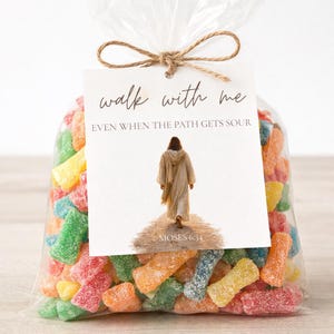 Op de afbeelding: Een transparante cellofaan zak gevuld met kleurrijke, zure snoepjes. Een label vastgebonden met touw leest "walk with me, even when the path gets sour" met een afbeelding van een persoon die op een pad loopt. De tekst "Moses 6:34" staat eronder.