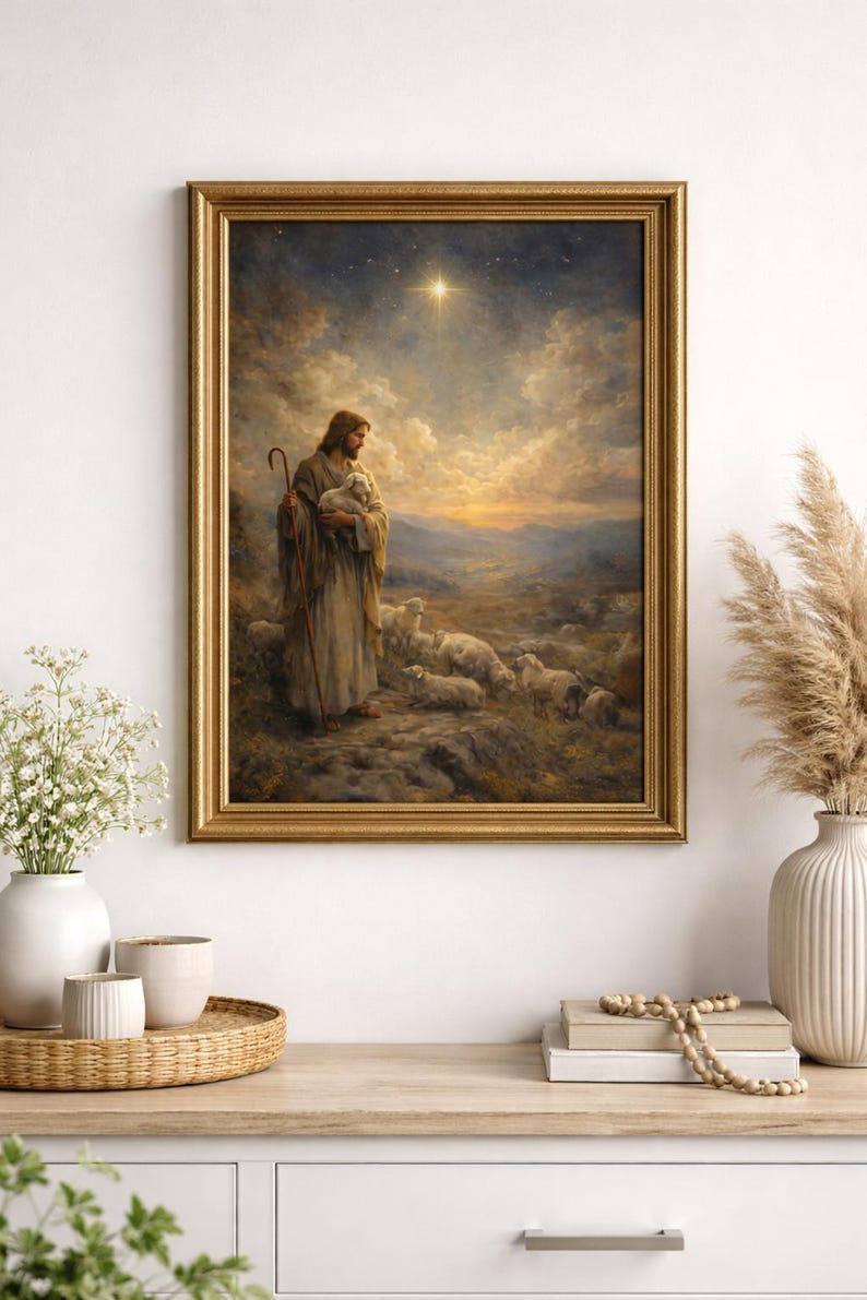 May include: A framed print depicts a religious scene with a figure holding a lamb, surrounded by sheep. The artwork is set against a backdrop of a starry sky and rolling hills, framed in a gold-colored ornate frame. The print is displayed on a white cabinet.