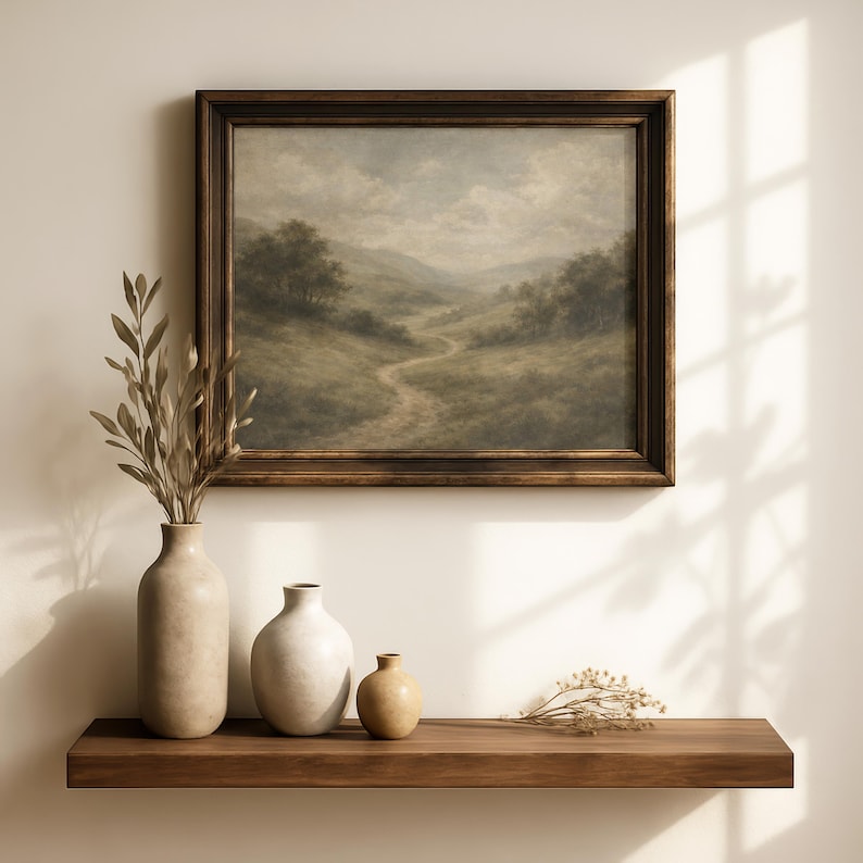 Pode incluir: Uma pintura de paisagem emoldurada com um caminho sinuoso atrav&eacute;s de um vale, exibida acima de uma prateleira de madeira. A pintura tem uma moldura de madeira escura. Tr&ecirc;s vasos de cer&acirc;mica e folhagens secas est&atilde;o na prateleira.
