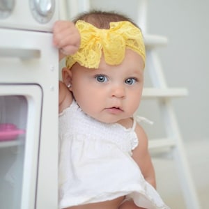 May include: A baby girl wearing a white dress and a yellow lace headband with a bow. The baby is looking at the camera and has blue eyes.