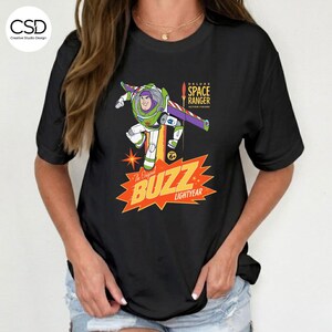 May include: Black t-shirt featuring a colorful graphic of Buzz Lightyear in a dynamic pose. The design includes the text "Deluxe Space Ranger Action Figure" and "The Original Buzz Lightyear" in a retro style. The shirt has short sleeves.