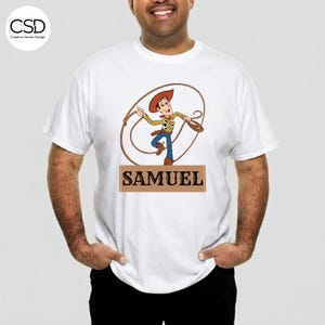 Puede incluir: Camiseta blanca con una imagen de dibujos animados de Woody de Toy Story, sosteniendo un lazo. El nombre "SAMUEL" est&aacute; impreso en una pancarta rectangular marr&oacute;n debajo. La camiseta es de cuello redondo.