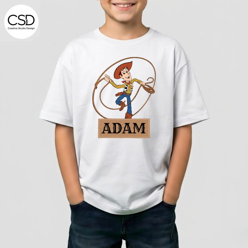 May include: White t-shirt featuring a cartoon cowboy character with a lasso. The character is wearing a cowboy hat, yellow shirt, and blue pants. The name "ADAM" is printed on a brown rectangle below the character.