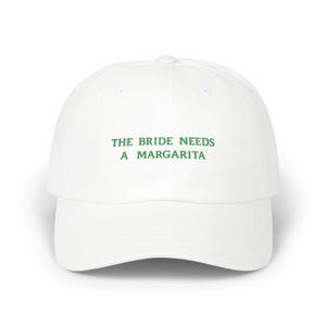 May include: White baseball cap with a curved brim. The cap features the text "THE BRIDE NEEDS A MARGARITA" in green, centered on the front. The hat is a classic, casual style.