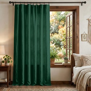 May include: A floor-length, solid dark green curtain panel hangs from a dark metal rod with rings. The fabric has a visible weave texture and vertical folds, covering a window frame that looks out onto a blurred outdoor garden scene.