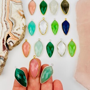 May include: A collection of marquise-shaped gemstone pendants with faceted surfaces and metallic edges in various colors including pink, green, white, blue, and dark gray, arranged in rows with some held in a hand for display.