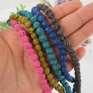 Op de afbeelding: Een close-up van zes strengen lavastenen kralen in verschillende kleuren, waaronder roze, geel, teal, blauw en grijs. De kralen zijn aan elkaar geregen en liggen op een hand.