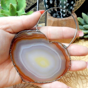 Puede incluir: Una rebanada de ágata en forma de corazón con un centro blanco y amarillo y bandas marrones alrededor del borde. El ágata está enmarcada con un lazo de alambre plateado.